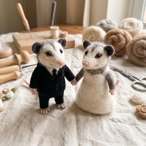 May include: Two needle-felted opossum figures standing side-by-side. The left opossum wears a black suit with a tie, while the right opossum wears a white dress with straps. Both figures have white and grey fur, dark eyes, and pink noses.