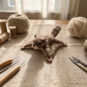 May include: A needle-felted flying squirrel figurine in shades of brown and cream with dark eyes, positioned on a textured fabric surface surrounded by wooden spools, balls of yarn, felting needles, and metal scissors for craft projects.