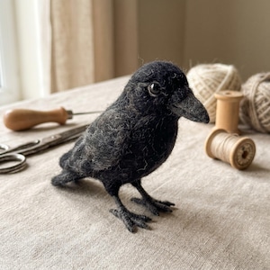 Op de afbeelding: Een naaldgevilt zwart kraai-figuurtje met een getextureerd wollen oppervlak, een spitse snavel en donkere glanzende ogen. De vogel staat op dunne, donkere, beweegbare pootjes en toont gedetailleerd vakmanschap in het zachte, donkere verenkleed.
