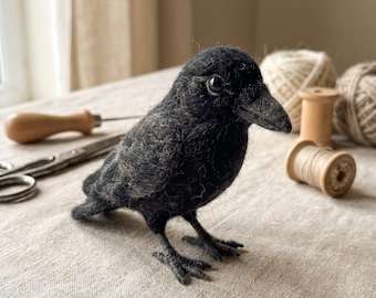May include: A needle-felted black crow figurine with a textured wool surface, a pointed beak, and dark glossy eyes. The bird stands on thin, dark, articulated legs, showcasing detailed craftsmanship in its soft, fuzzy, and dark plumage.
