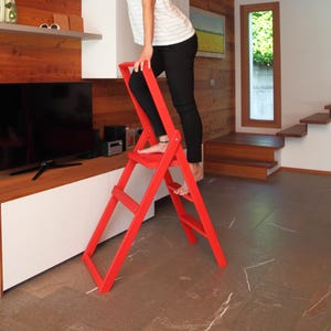 Puede incluir: Una escalera de mano plegable de color rojo brillante con tres peldaños, construida con un marco sólido. La escalera cuenta con un asa alta en la parte superior para mayor estabilidad y apoyo, con un diseño articulado para plegarla.