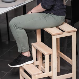 Könnte beinhalten: Ein heller Holztritt mit zweistufigem Design, bestehend aus einer höheren Sitzfläche und einer unteren Stufe. Der Hocker ist aus Naturholz mit sichtbarer Maserung gefertigt und verfügt über vier stabile Beine sowie horizontale Sitzlatten.