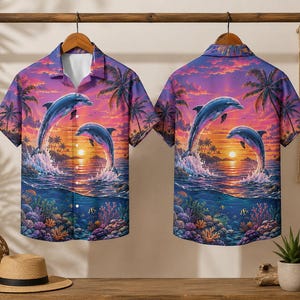 Könnte beinhalten: Kurzärmeliges Hemd mit tropischem Sonnenuntergangsmotiv. Es zeigt springende Delfine, Palmen und Korallen vor einem Himmel mit Farbverlauf in Lila, Orange und Blau. Das Design wiederholt sich auf Vorder- und Rückseite.