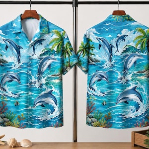 Könnte beinhalten: Ein kurzärmeliges Button-Down-Hemd mit einer lebendigen blauen Ozeanszene mit springenden Delfinen, Palmen und Korallen. Das Design des Hemdes wiederholt sich auf der Vorder- und Rückseite und schafft ein zusammenhängendes tropisches Thema. Das Hemd hängt an einem Metallgestell.