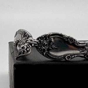 May include: A silver-toned metal bracelet crafted from a repurposed spoon handle featuring an ornate floral embossed pattern. A small, intricate heart-shaped charm with a filigree design is attached to the band by a circular metal jump ring.