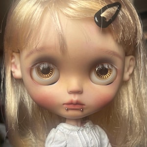 Pode incluir: Um close-up do rosto de uma boneca com cabelo loiro e olhos grandes castanho-dourados. A boneca tem uma tez pálida com bochechas rosadas e um pequeno piercing labial prateado. Uma presilha preta está no cabelo da boneca. A boneca está vestindo uma blusa branca.