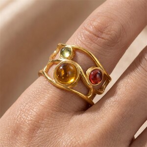 May include: A gold-toned ring featuring an open, organic, branch-like band design. The ring is set with three round, smooth, cabochon-style stones: one large amber-colored stone, one smaller olive-green stone, and one smaller deep red stone.