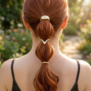 Può includere: Un set di tre accessori per capelli color oro disposti su una coda di cavallo. Il pezzo superiore è una cupola liscia e arrotondata, mentre i pezzi centrale e inferiore sono polsini metallici a forma di V fissati lungo la lunghezza dei capelli.