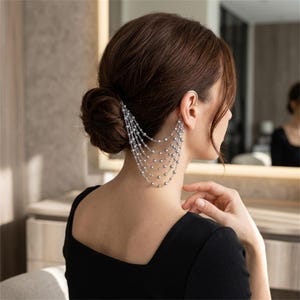 Puede incluir: Un pendiente tipo ear cuff de tono plateado conectado a una serie de seis cadenas plateadas en cascada adornadas con pequeñas perlas blancas redondas. La pieza de joyería se sujeta al cartílago superior de la oreja y cae hacia la nuca.