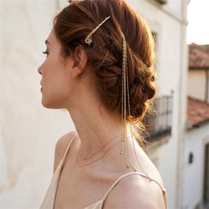 Puede incluir: Un accesorio para el cabello en tono dorado que presenta una base de horquilla con múltiples cadenas delgadas y delicadas que cuelgan. Las cadenas son metálicas y crean un efecto de capas sobre el cabello, sujetas por un pasador decorativo.