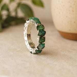 May include: A silver ring featuring a series of raw, uncut green emerald stones set along the band. The metal band has a textured, organic finish that complements the irregular shapes and deep green hues of the natural gemstone pieces.