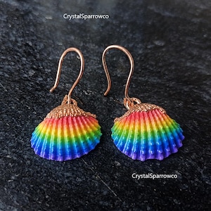 Può includere: Un paio di orecchini pendenti con ciondoli a forma di conchiglia naturale dipinti con un vivace gradiente arcobaleno di rosso, arancione, giallo, verde, blu e viola. Le conchiglie sono fissate a ganci in metallo color oro rosa. Testo CrystalSparrowco.