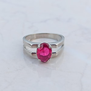May include: A silver-toned ring featuring a central oval-cut vibrant pink gemstone held by four prongs. The band has a split-shank design that tapers toward the back, creating a clean and polished look for this piece of jewelry.