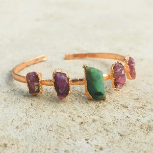 May include: A copper-toned cuff bracelet featuring a series of five raw, uncut gemstones. The stones include four deep pink crystals and one central green crystal, each set in a textured metal bezel along the open-ended band design.
