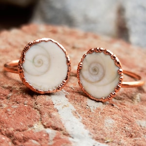 Può includere: Due anelli con cabochon circolari in conchiglia occhio di shiva color bianco sporco, montati su fasce in metallo testurizzato color rame. Ogni conchiglia presenta un motivo a spirale naturale al centro, incorniciato da una montatura smerlata.