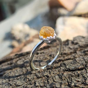 May include: A silver ring featuring a single, raw, amber-coloured gemstone set in a textured, crown-like prong setting. The band is smooth and polished, contrasting with the rough, irregular surface of the central stone and the detailed metalwork.