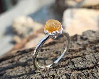 May include: A silver ring featuring a single, raw, amber-colored gemstone set in a textured, crown-like prong setting. The band is smooth and polished, contrasting with the rough, irregular surface of the central stone and the detailed metalwork.