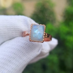 Può includere: Un anello con una pietra di luna rettangolare sfaccettata montata su una fascia in metallo color rame. La pietra mostra un bagliore blu traslucido e iridescente. L'incastonatura presenta un bordo decorativo a perline che incornicia la gemma.