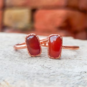 Puede incluir: Dos anillos con piedras rectangulares de color rojo intenso y translúcido engastadas en bandas de cobre texturizado. Cada piedra está asegurada por un engaste de metal festoneado, mostrando una superficie lisa y pulida que contrasta con el cobre martillado.