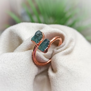May include: A copper-toned ring featuring two raw, uncut teal-colored stones set side-by-side. The metal band has a smooth, polished finish and an open design, showcasing the natural, irregular texture of the two blue-green mineral crystals.