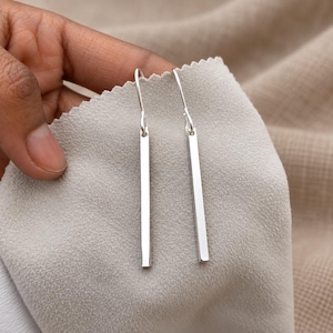May include: A pair of silver-toned drop earrings featuring long, slender, rectangular bar pendants suspended from simple curved ear wires. The metal surface is smooth and reflective, showcasing a clean, minimalist design with a polished finish.
