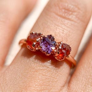 May include: A rose gold ring featuring three raw, uncut gemstones set in a horizontal row. The centre stone is a deep purple, while the two flanking stones are a vibrant, translucent orange. The metal band is smooth and polished in a rose gold tone.