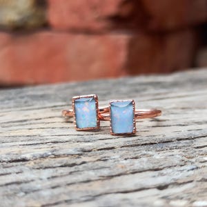 May include: Two copper rings featuring rectangular, light blue opalescent stones with iridescent flashes of colour. The metal bands have a textured, rustic finish, and the stone settings are framed by a delicate, scalloped metal edge detail.