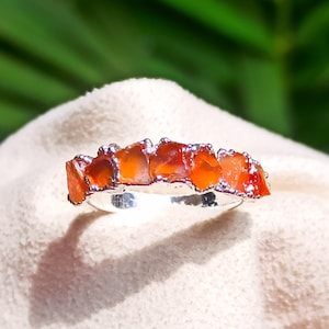 May include: A silver ring featuring a row of five raw, vibrant orange carnelian stones set along the top. The band is polished silver, and the stones have irregular, jagged edges, creating a textured appearance across the top of the ring.