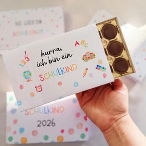 Pode incluir: Uma caixa de chocolates retangular branca com uma bandeja dourada contendo três chocolates redondos. A caixa apresenta ilustrações coloridas de um globo, despertador, escola, lápis, tintas e o texto: hurra, ich bin ein SCHULKIND.