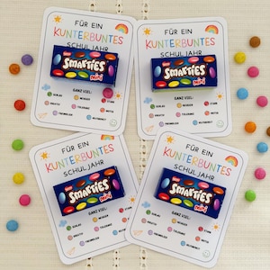 Könnte beinhalten: Vier rechteckige weiße Karten mit abgerundeten Ecken, auf denen jeweils eine kleine blaue Schachtel Smarties mini befestigt ist. Die Karten zeigen den Text FÜR EIN KUNTERBUNTES SCHULJAHR und GANZ VIEL mit bunten Symbolen und deutschen Wörtern.