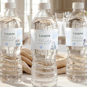 Peut inclure: Trois bouteilles d'eau en plastique transparent avec des bouchons blancs, chacune avec une étiquette personnalisée portant le texte Louane Baby Shower, des accents bleu clair, un flocon, un cœur bleu et des illustrations d'animaux.