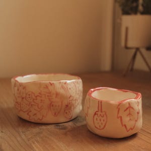 Puede incluir: Dos cuencos de cerámica con diseño de fresas. Los cuencos son de color blanco roto con un patrón de fresas rojas en los laterales. El cuenco más grande tiene unos 13 cm de diámetro y el más pequeño unos 8 cm.
