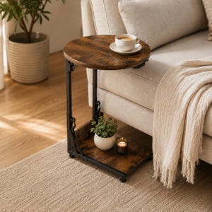 May include: A C-shaped side table with a round dark brown wood-grain top and a matching rectangular base connected by a black metal frame. The table holds a white ceramic cup on a saucer, a small potted green plant, and a glass candle holder.