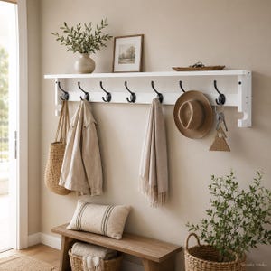 Op de afbeelding: Een witte wandkapstok met zeven zwarte metalen haken. De bovenste plank bevat een vaas met groen, een fotolijst en een klein dienblad. Aan de haken hangen een gevlochten tas, een beige jas, een sjaal en een hoed.