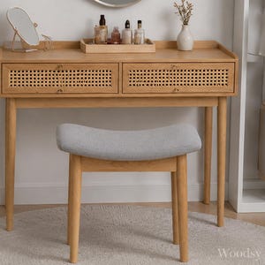 May include: A light wood vanity table featuring two drawers with woven cane fronts and small gold knobs. The table holds a small oval mirror, a wooden tray with various bottles, and a white vase. A matching light wood stool with a grey upholstered seat sits in front.