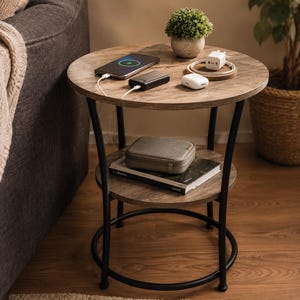 May include: A round side table with a wood-grain finish top and a lower shelf, supported by a black metal frame. The top surface holds a smartphone, a power bank, a white charging block, and a small potted plant. A gray case sits on a book below.
