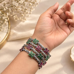 May include: A set of four silver-toned metal bangles featuring a textured, oxidised finish. Each bangle is adorned with multiple four-petaled flower motifs, alternating between vibrant green and deep pink gemstone-like inlays throughout.