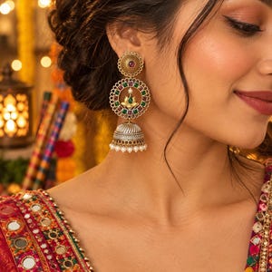 May include: A gold-toned jhumka earring featuring a circular stud with a central red stone, a larger circular frame adorned with alternating green and red square stones surrounding a central lotus motif, and a silver-toned bell-shaped base with white beads.