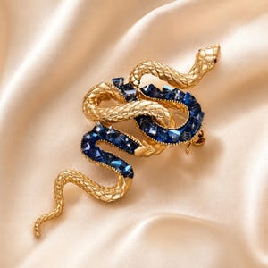 Pode incluir: Um broche de serpente em tom dourado com corpo texturizado e escamado, apresentando uma seção enrolada incrustada com cristais facetados azul-escuros. A cabeça da serpente está elevada com pequenos olhos vermelhos e acabamento polido.