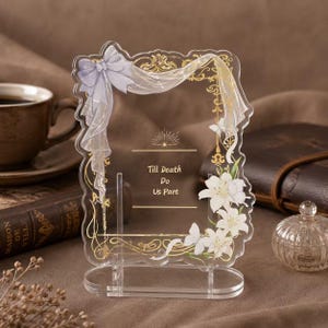Op de afbeelding: Een rechthoekig bord van helder acryl met een geschulpte rand, voorzien van een gouden filigraanrand, een lichtpaarse strik en witte lelie-illustraties. De tekst in het midden luidt "Till Death Do Us Part" in gouden letters.