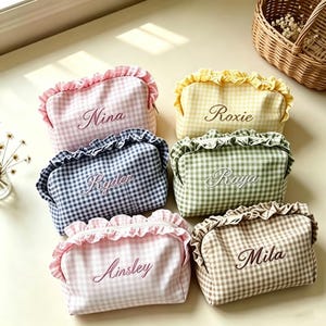 Pode incluir: Seis estojos retangulares com zíper, bordas com babados e padrões xadrez vichy em rosa, amarelo, azul-marinho, verde, rosa claro e bege. Cada estojo apresenta os nomes bordados Nina, Roxie, Ryder, Raya, Ainsley e Mila em letra cursiva.
