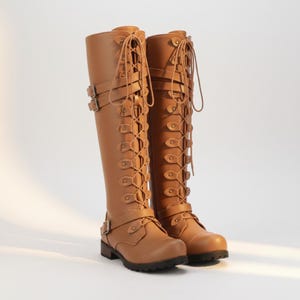 May include: A pair of knee-high tan boots featuring a full-length front lace-up design with metal eyelets, two adjustable buckled straps across the upper shaft, and a single buckled strap at the ankle. The boots have a rounded toe and a low heel.