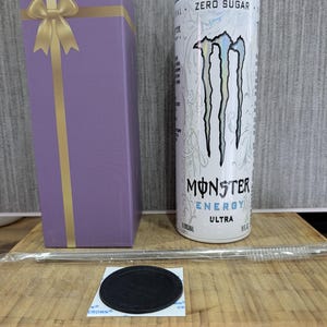 Könnte beinhalten: Eine lila Geschenkbox mit goldener Schleife, eine weiß-silberne Monster Energy Ultra Dose, ein Edelstahlstrohhalm und ein schwarzer Untersetzer. Die Dose zeigt das Monster-Logo und die Worte "ZERO SUGAR".