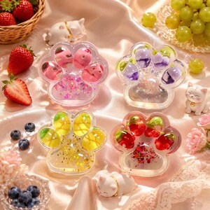 Op de afbeelding: Vier doorzichtige onderzetters van hars in de vorm van een poot, gevuld met kleurrijke fruitvormige inzetstukken en glitters, vergezeld van kleine witte kattenbeeldjes, verse aardbeien, bosbessen en groene druiven op een gedrapeerde stof.
