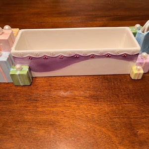 May include: A ceramic dish shaped like a row of presents with a purple and white design. The dish has a white interior and is decorated with pearl-like accents.