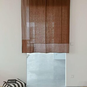 May include: A rectangular brown curtain with a visible woven texture, featuring a darker brown panel along the bottom edge. The curtain is suspended from a thin black rod with circular rings, covering the upper portion of a doorway opening.