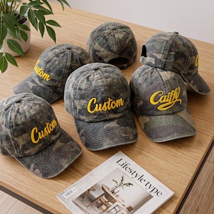 May include: A collection of camouflage baseball caps featuring embroidered yellow text. Several caps display the word Custom in a cursive font, while one cap features the word Caiffy in yellow script. Each cap has a curved brim and eyelets.