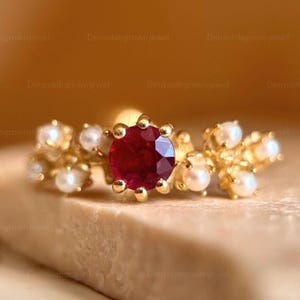 Può includere: Un anello in oro con un rubino rosso intenso a taglio rotondo al centro, circondato da un gruppo di piccole perle bianche su ciascun lato. La fascia è in oro giallo e il design presenta una disposizione simmetrica delle perle e del rubino.