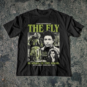 Puede incluir: Una camiseta negra de manga corta con un estampado gráfico de la película La Mosca de 1986. El diseño incluye el título, una criatura, una cabina de laboratorio y retratos de Jeff Goldblum y Geena Davis con el texto Be Afraid. Be Very Afraid.
