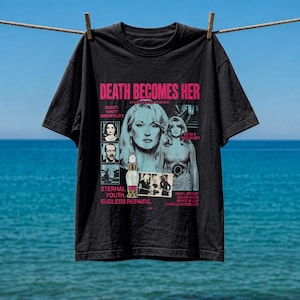 Puede incluir: Una camiseta negra de manga corta con un estampado gráfico de la película Death Becomes Her. El diseño incluye texto en rosa, retratos del reparto, una ilustración de un frasco de poción y las frases Beauty, Vanity, Immortality y Eternal Youth.